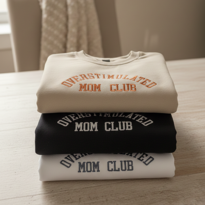 Stack of sweatshirts with 'Overstimulated Mom Club' text on a wooden surface.